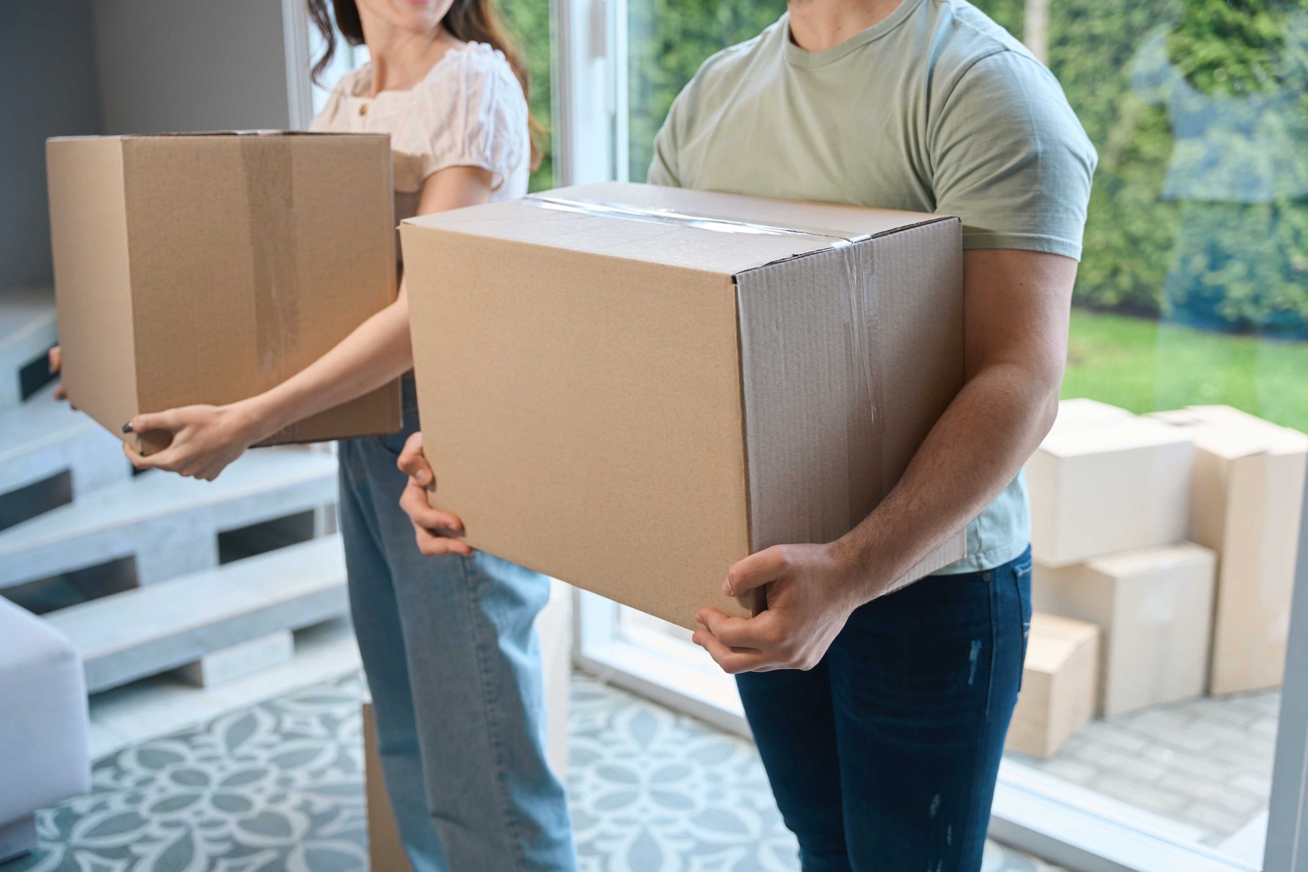 Two people carry large cardboard boxes indoors, with more boxes in the background, suggesting a move—perhaps with the help of Long Distance Movers Chicago. Their faces are not visible in the image.