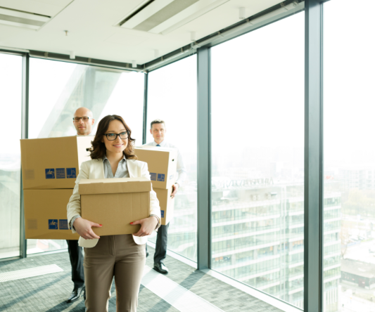 Three people in business attire carry cardboard boxes in a modern office with large windows and a city view, suggesting they are moving with the help of Long Distance Movers Chicago IL.