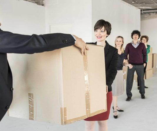 Five people stand in a line in an unfinished office space, passing large cardboard boxes to each other—just like experienced Long Distance Movers Chicago organizing a big move, all dressed in business attire.