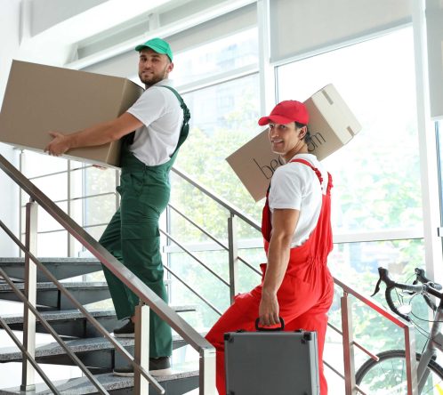 Two movers in colored overalls and caps, from Long Distance Movers Chicago, carry cardboard boxes and a toolbox up a staircase in a bright, modern building. A bicycle is visible near the window in the background.