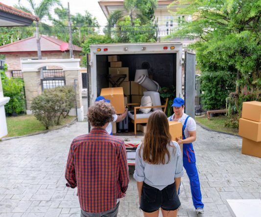 Two people stand outside their house in a suburban neighborhood, watching Long Distance Movers Chicago unload furniture and boxes from a moving truck parked in the driveway, surrounded by greenery and nearby homes.