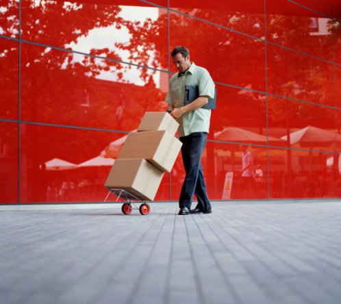 A man pushes a hand truck loaded with cardboard boxes along a sidewalk, passing by a modern building with a bright red, reflective glass facade—just another busy day for Long Distance Movers in Chicago.