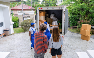 Two people stand outside, watching as Long Distance Movers Chicago load a truck with furniture, boxes, and plants in a driveway surrounded by greenery and houses.