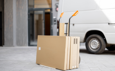 A hand truck with cardboard boxes stacked on it stands on a paved surface next to a white van, in front of a building entrance—just the scene you’d expect with Long Distance Movers Chicago handling your relocation.