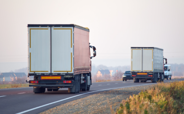 Two large box trucks from Long Distance Movers Chicago drive on a rural road, with cars visible ahead. It’s daylight, and houses and fields make up the backdrop as both trucks travel in the same direction.