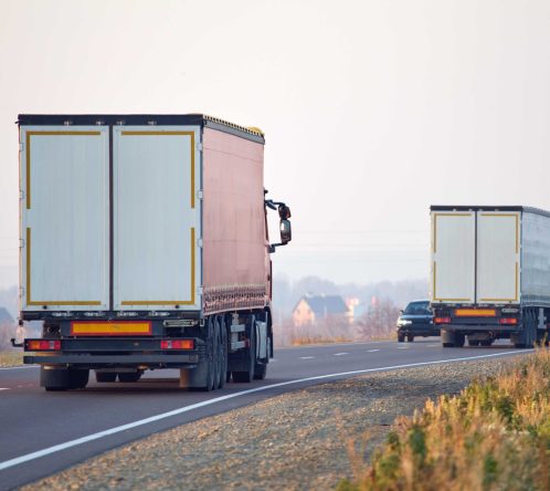 Two large box trucks from Long Distance Movers Chicago drive along a rural road, with two cars following behind. The scene is set in daylight, with grassy shoulders and distant houses visible in the background.